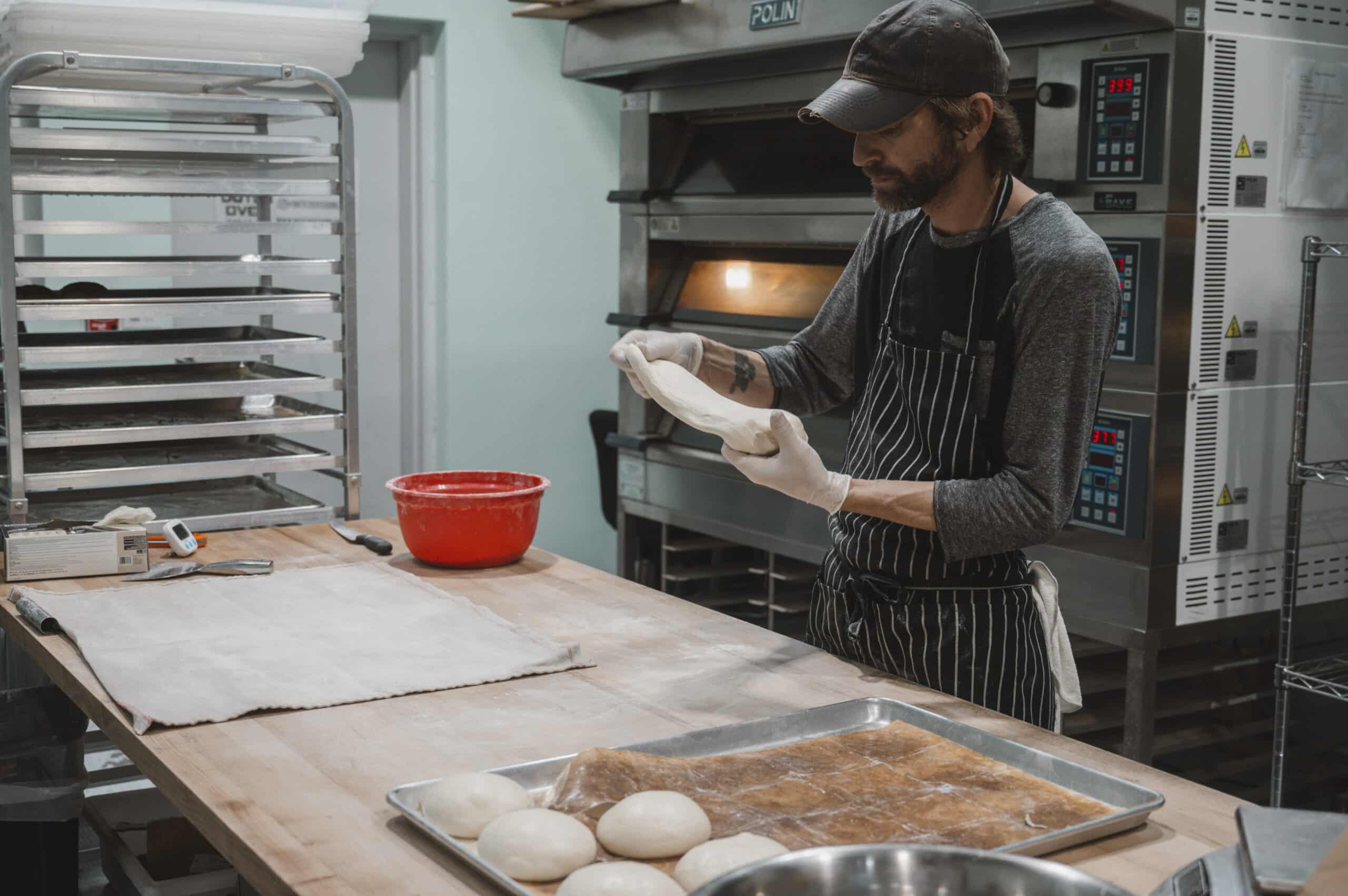 Baker shaping dough in a bakery with oven and proofing rack, preparing sourdough bread.