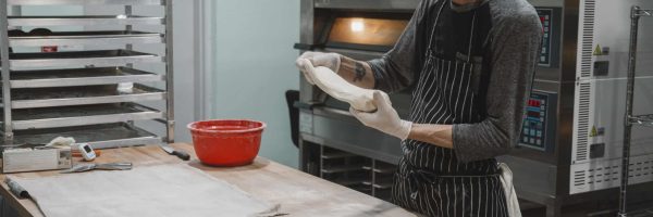 Baker shaping dough in a bakery with oven and proofing rack, preparing sourdough bread.